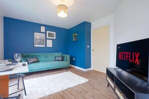 A living room with a teal sofa, a white rug, a TV with "NETFLIX" on it, and a wall with framed pictures.