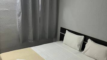 A hotel room with a bed, white linens, a beige bedspread, a white pillow, a white towel, and a window with a curtain.