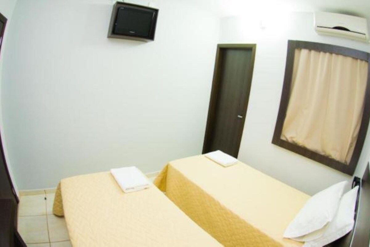 Une petite chambre avec un lit simple, un téléviseur fixé au mur, une porte et une fenêtre avec des rideaux.