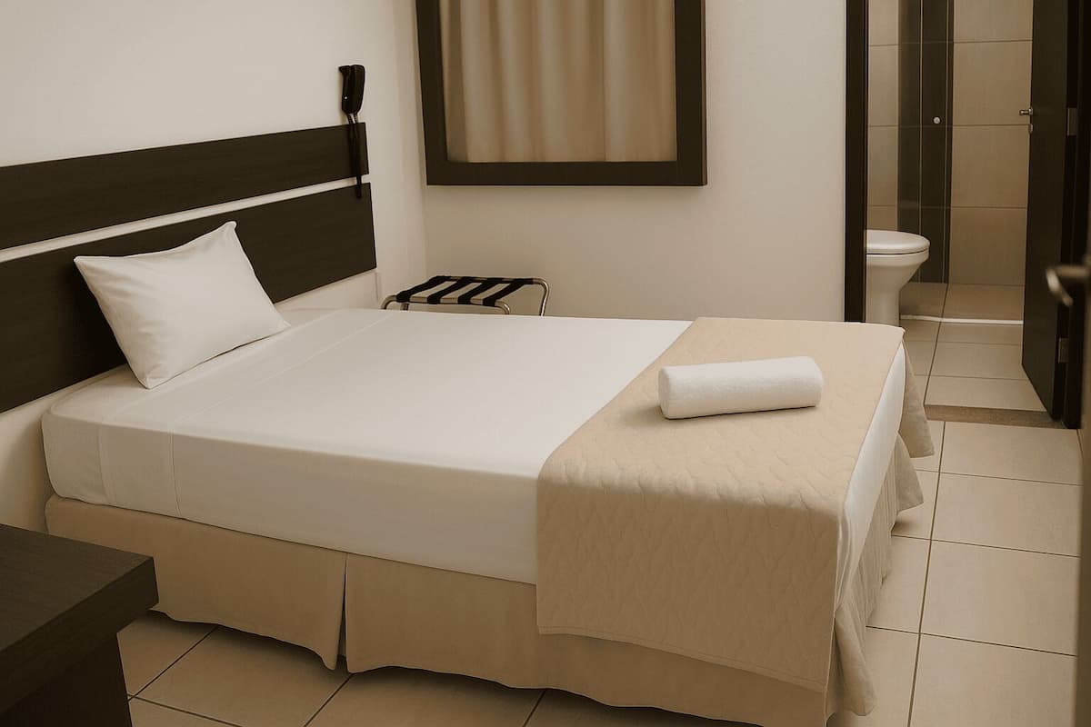 A neatly made bed with a white comforter and a beige blanket, a bedside table with a lamp, and a bathroom visible through an open door.