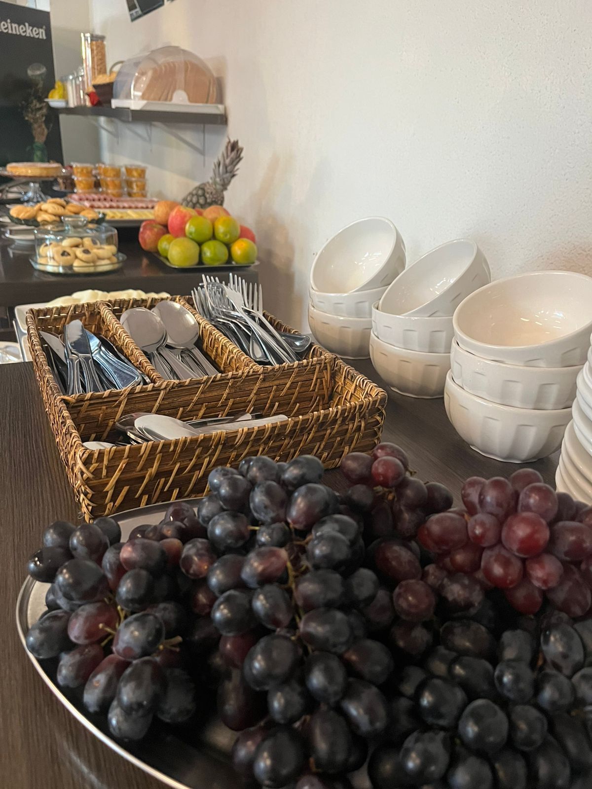 Una mesa de buffet con una cesta de mimbre que contiene utensilios, un bol de uvas y una bandeja con frutas y pasteles.
