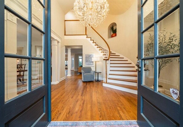 Un intérieur spacieux avec un escalier en bois, un lustre et une vue dégagée sur l’extérieur grâce à des portes vitrées.