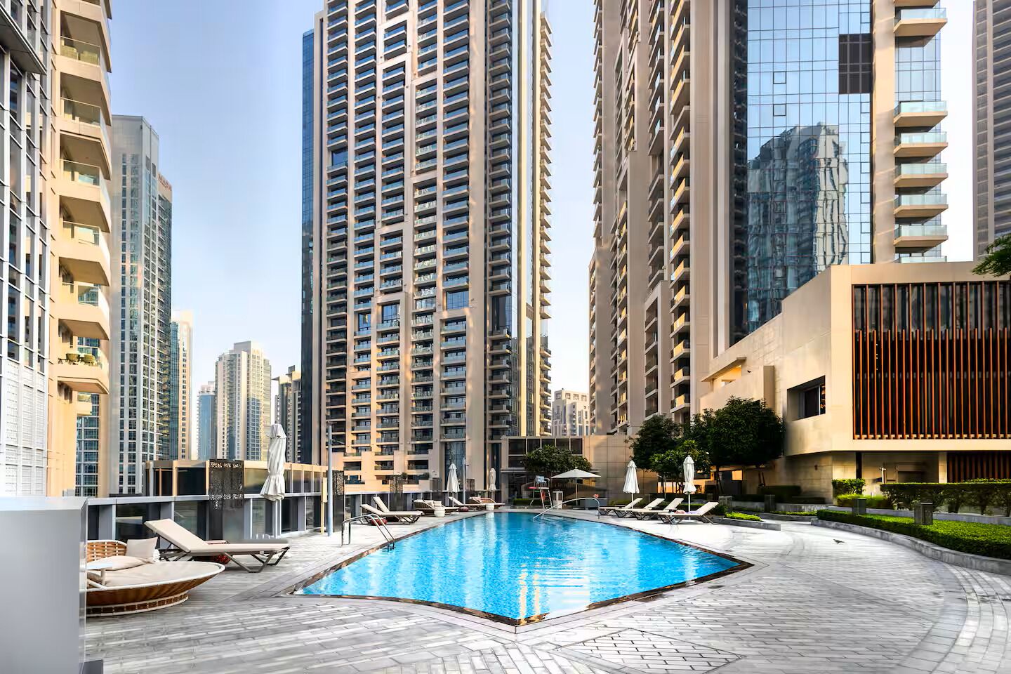 A modern residential complex with a central swimming pool surrounded by lounge chairs and sunbeds.