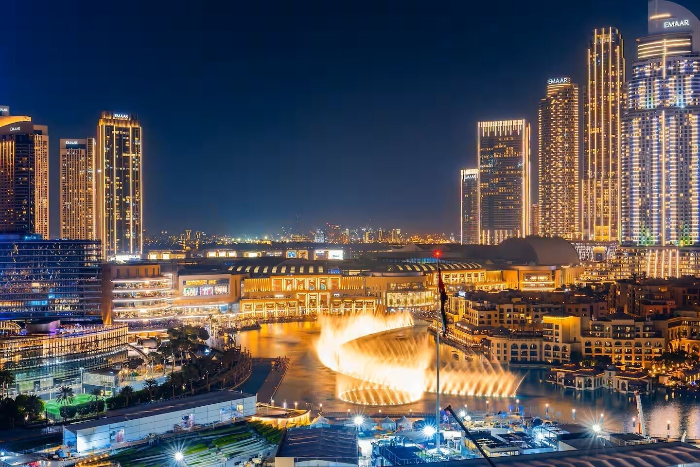 A cityscape at night with illuminated buildings and a central fountain.