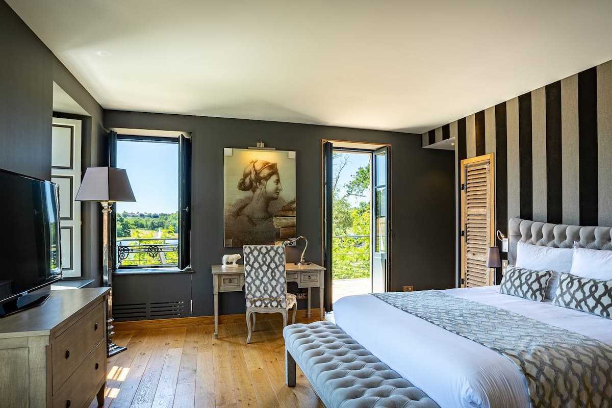 A bedroom with a large bed, a desk, a chair, a television, and a window with a view of greenery.
