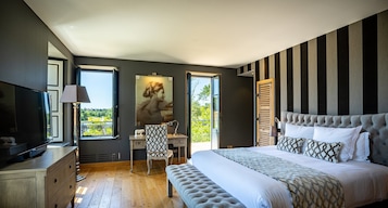 A bedroom with a large bed, a desk, a chair, a television, and a window with a view of greenery.