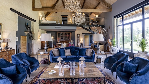 Amplia sala de estar con un sofá azul grande, dos sillones y una mesa de centro con velas. Un candelabro cuelga del techo, y hay una escalera que conduce a un nivel superior.