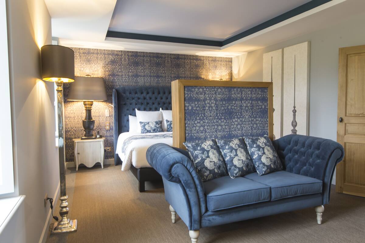 A bedroom with a bed, a blue sofa, a nightstand with a lamp, and a patterned wall.
