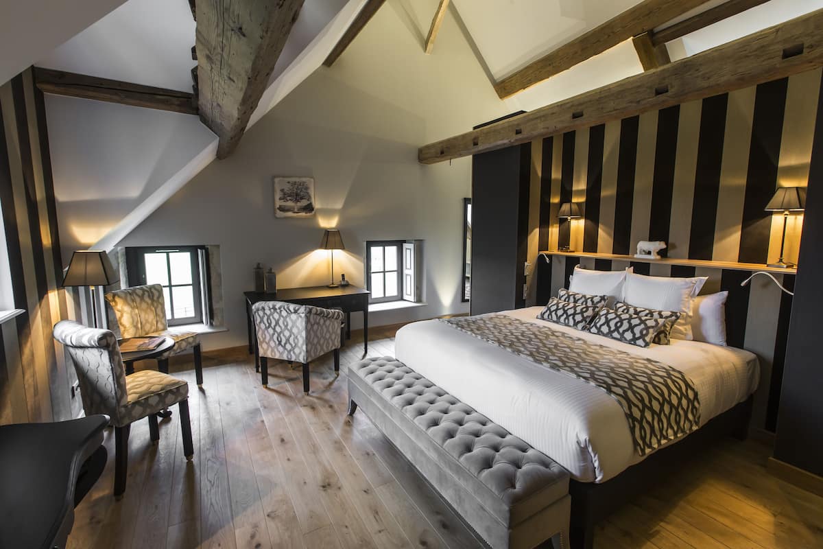 A bedroom with a large bed, a bench, a desk, and a chair. There are windows and a wooden ceiling beam.