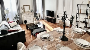 A dining area with a wooden table set for six, surrounded by a modern living room with a sofa, a TV, and decorative items.