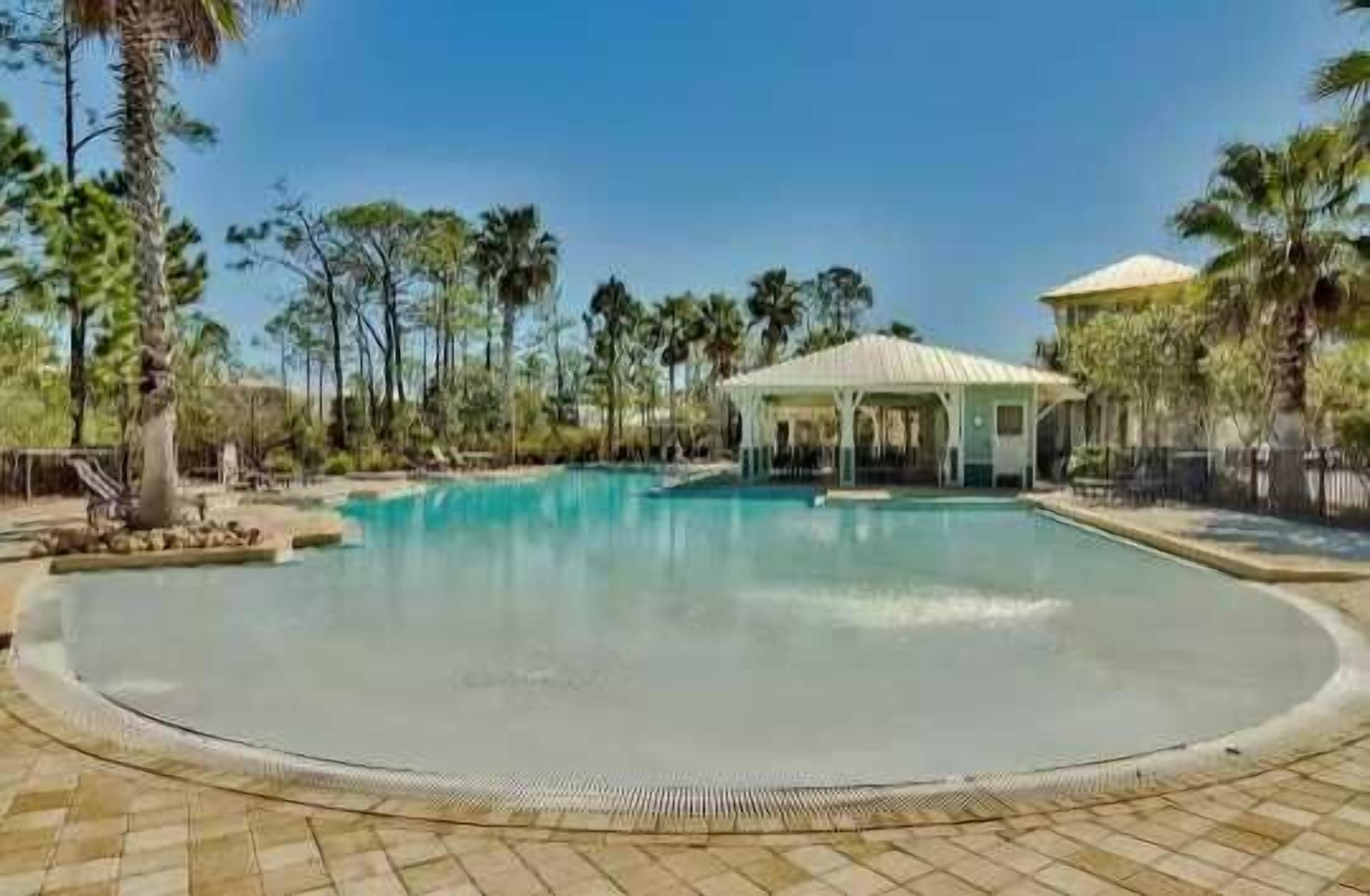 A large outdoor swimming pool with a covered pavilion, surrounded by palm trees and a clear sky.