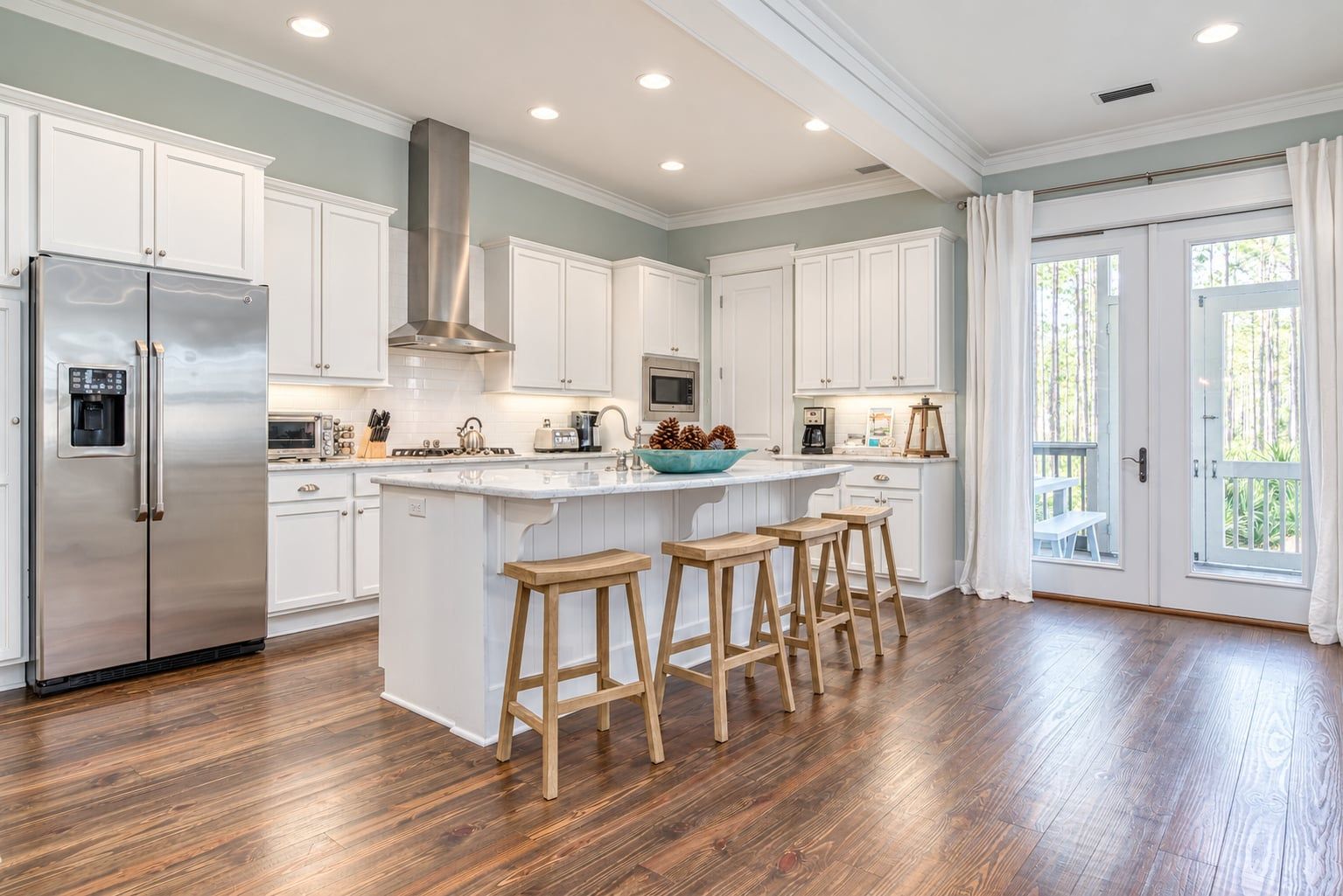 A spacious kitchen with white cabinetry, a stainless steel refrigerator, wooden stools, and a large island with a bowl of pinecones.