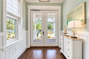 A hallway with a double door leading to an outdoor area, a white dresser with a lamp, and a framed picture on the wall.