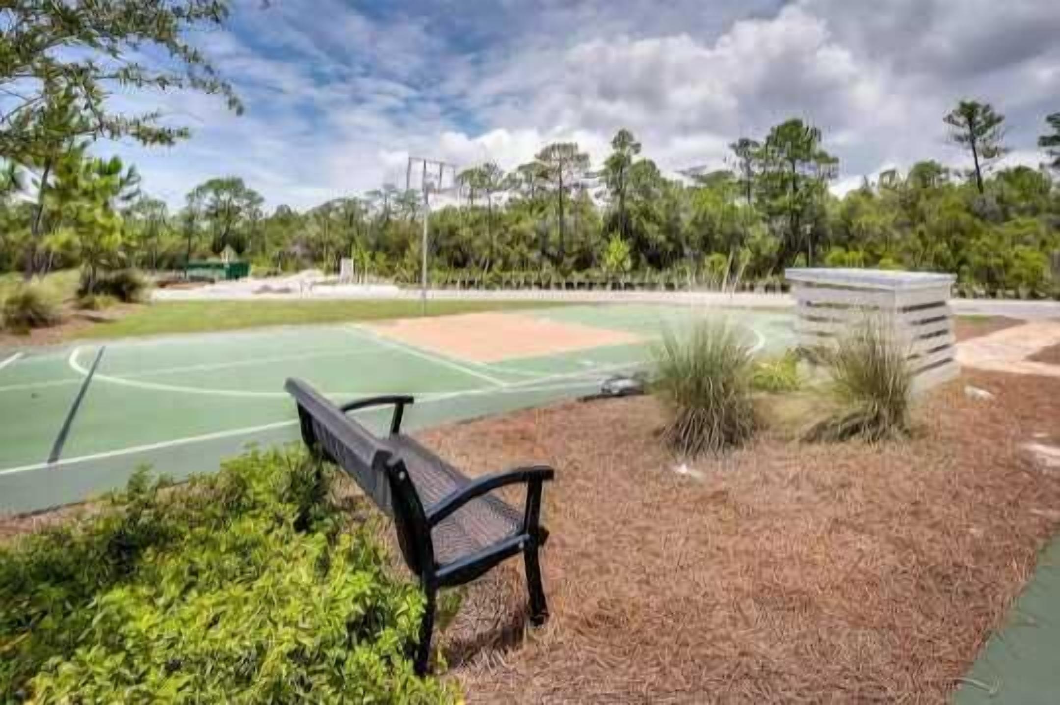 A tennis court with a bench, surrounded by trees and a grassy area.
