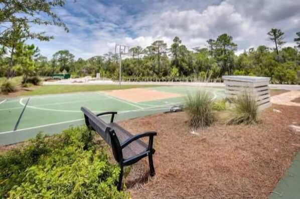 A tennis court with a bench, surrounded by trees and a grassy area. - Salty Days - 5BR/3BA NatureWalk Home - Sleeps 12 - Golf Cart & Community Pools (Santa Rosa Beach)