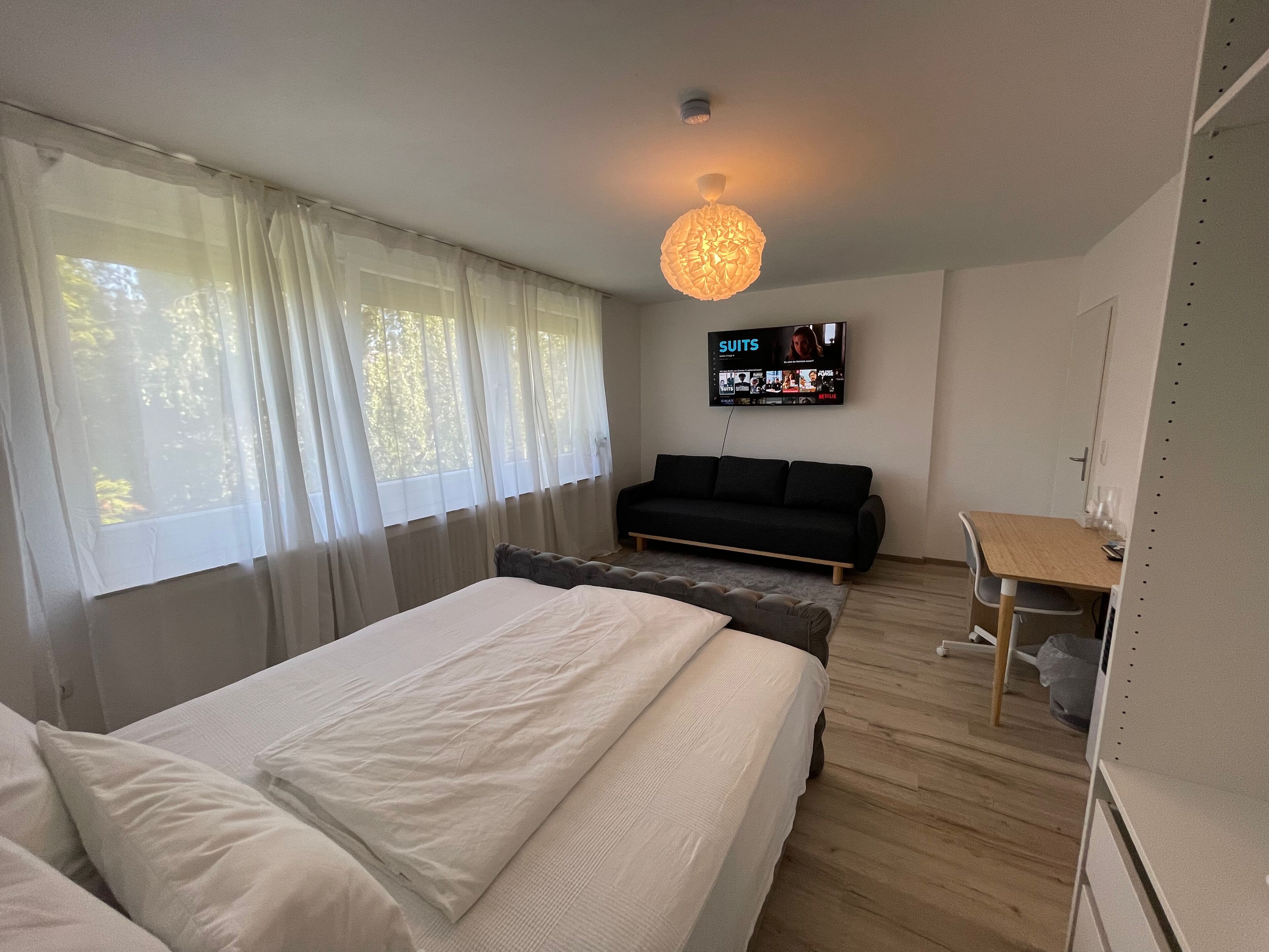 A modern hotel room with a large bed, a sofa, a TV mounted on the wall, and a wooden floor.