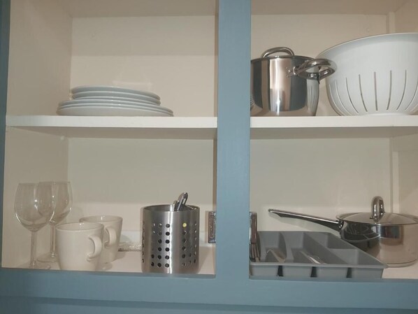Une cuisine avec des étagères blanches, où l’on trouve des assiettes, des tasses, une passoire, une casserole et une poêle.