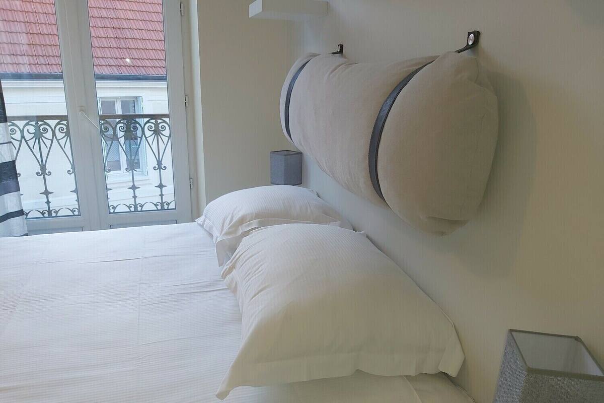 Un dormitorio con cama, almohadas, una mesita de noche y una ventana con vistas a un balcón.