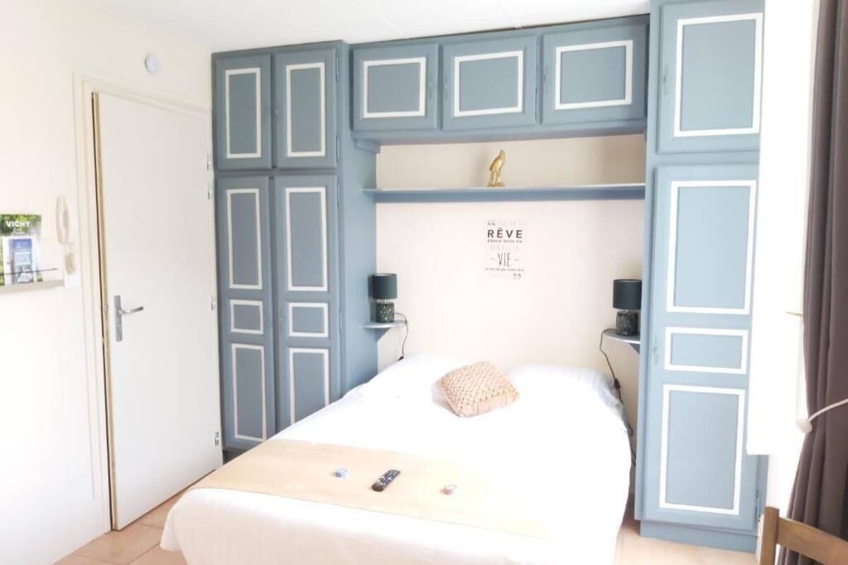 Un dormitorio con cama, mesita de noche, repisa en la pared con un objeto decorativo y una puerta.