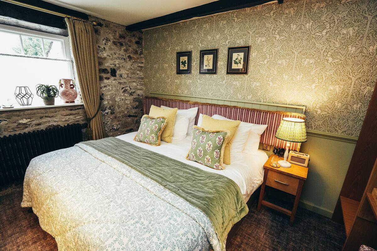 A bedroom with a stone wall, a bed with patterned bedding, a nightstand with a lamp, and three framed pictures on the wall.