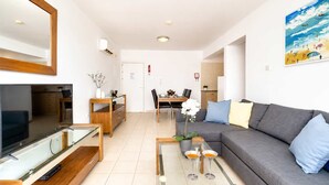 A modern living room with a flat-screen TV, a sofa, a coffee table, and a dining area with a table and chairs. - 2BR Apartment with Pool - Poppy by Premier (Paralimni)