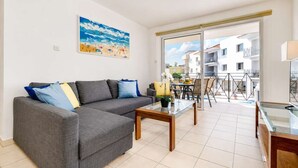 A modern living room with a grey sofa, a glass coffee table, and a painting of a beach scene. - 2BR Apartment with Pool - Poppy by Premier (Paralimni)