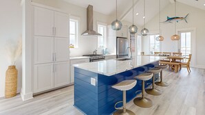 A modern kitchen with white cabinets, a blue island, hanging pendant lights, and a dining area with wooden furniture.