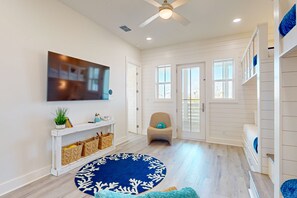 A spacious living room with a large flat-screen TV, a ceiling fan, and a blue patterned rug. - Private Heated Pool, Resort Pools, Golf Cart Boardwalk to the Beach (Port Aransas)