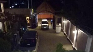 A nighttime view of a residential area with a paved driveway leading to a house. There is a car parked on the left and a person standing near the entrance. - BAVAH AIRPORT HOTEL (Entebbe)
