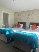 Habitación de hotel con dos camas, cada una con colcha color turquesa y almohadas naranjas, cabecero gris y papel tapiz con motivos florales.