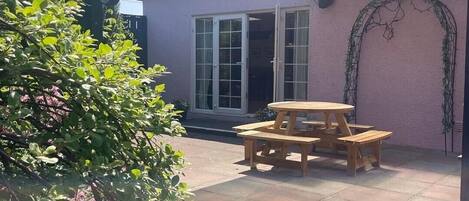 Un patio con mesa y bancos de madera, un área techada y un edificio con puerta y ventanas.