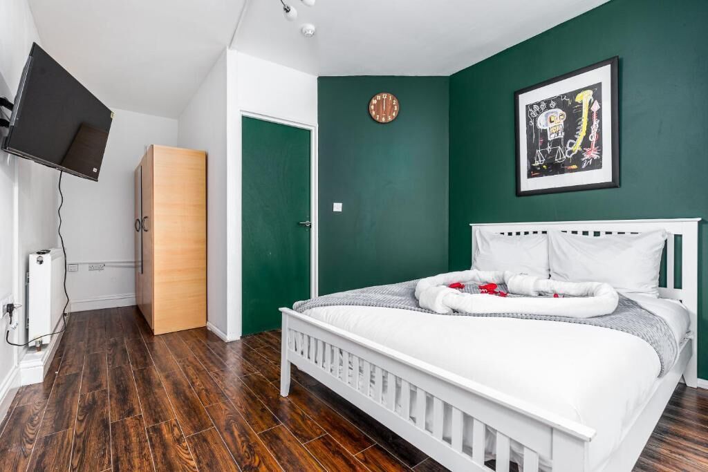 Une chambre à coucher avec un lit blanc, des murs verts, une armoire en bois, une horloge et un tableau encadré.