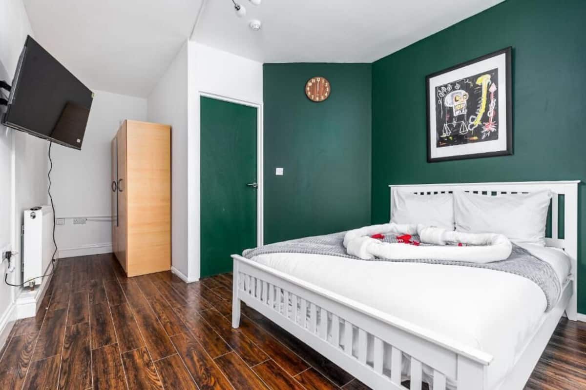 A bedroom with a white bed, green walls, a wooden wardrobe, a clock, and a framed picture.
