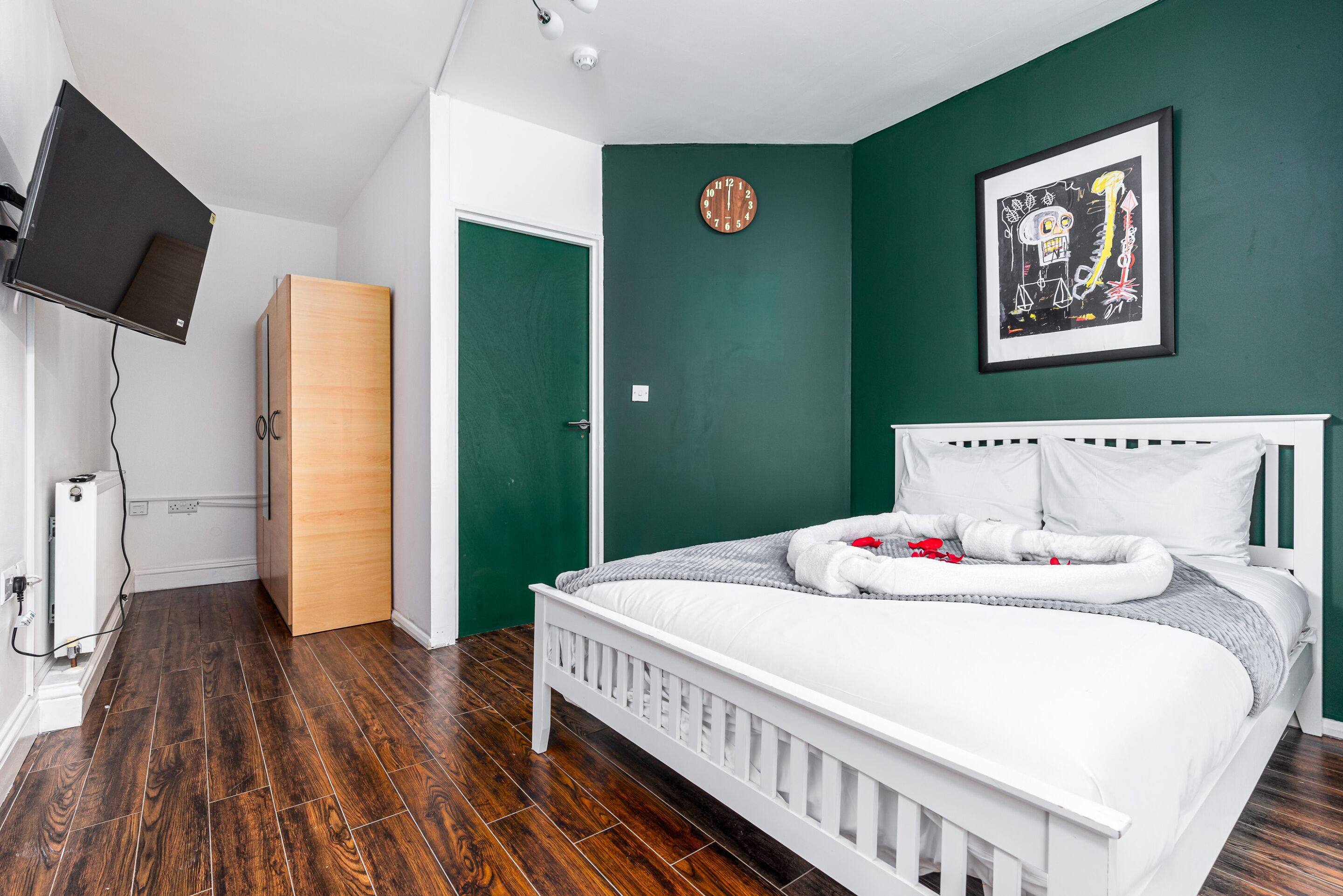 A bedroom with a white bed, green walls, a wooden wardrobe, a clock, and a framed artwork.