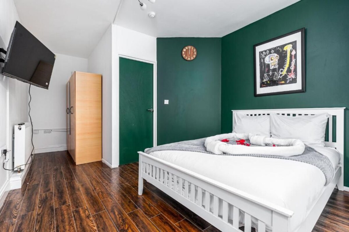 Quarto com cama branca, paredes verdes, guarda-roupa de madeira, relógio e quadro emoldurado.