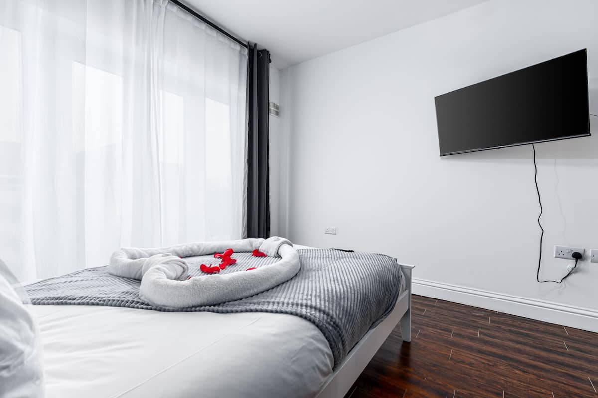 A modern hotel room with a bed, a flat-screen TV mounted on the wall, and a window with sheer curtains.