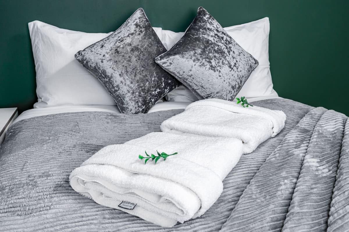 A neatly made bed with white pillows and a gray quilt, a white towel with a green leaf, and a green wall in the background.