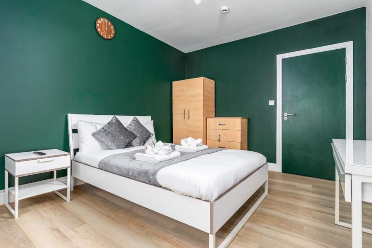 A bedroom with a bed, a wooden wardrobe, a white bedside table, and a green wall.