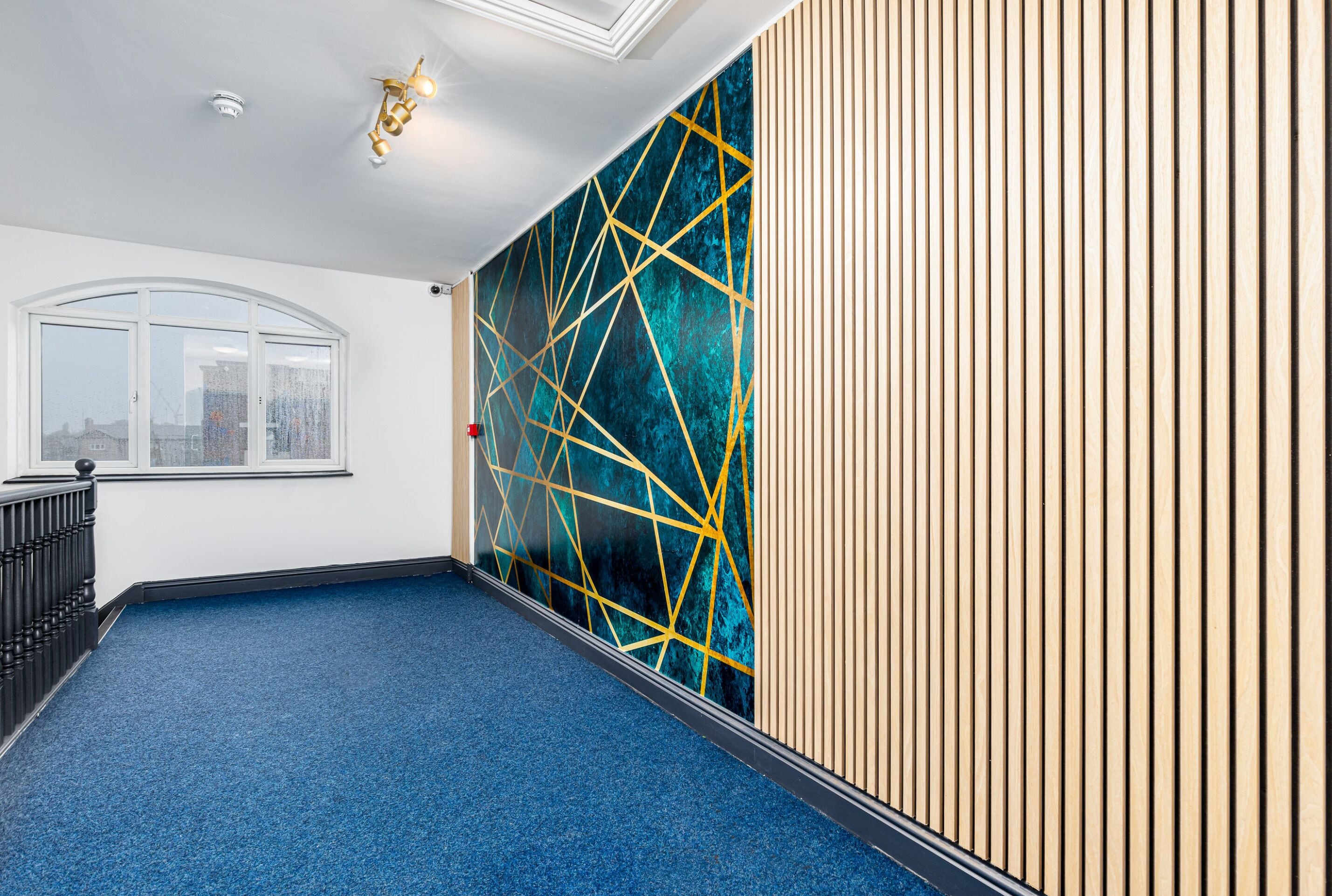Un pasillo con alfombra azul, una pared blanca con un ventanal, y una pared con listones de madera verticales y un diseño geométrico.