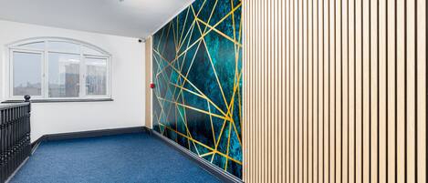 Un pasillo con alfombra azul, una pared blanca con un ventanal, y una pared con listones de madera verticales y un diseño geométrico.