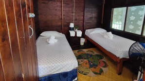 Ein Zimmer mit zwei Betten, einem Holzschrank, einem kleinen Tisch mit Lampe und einem Fenster mit floralem Vorhang.
