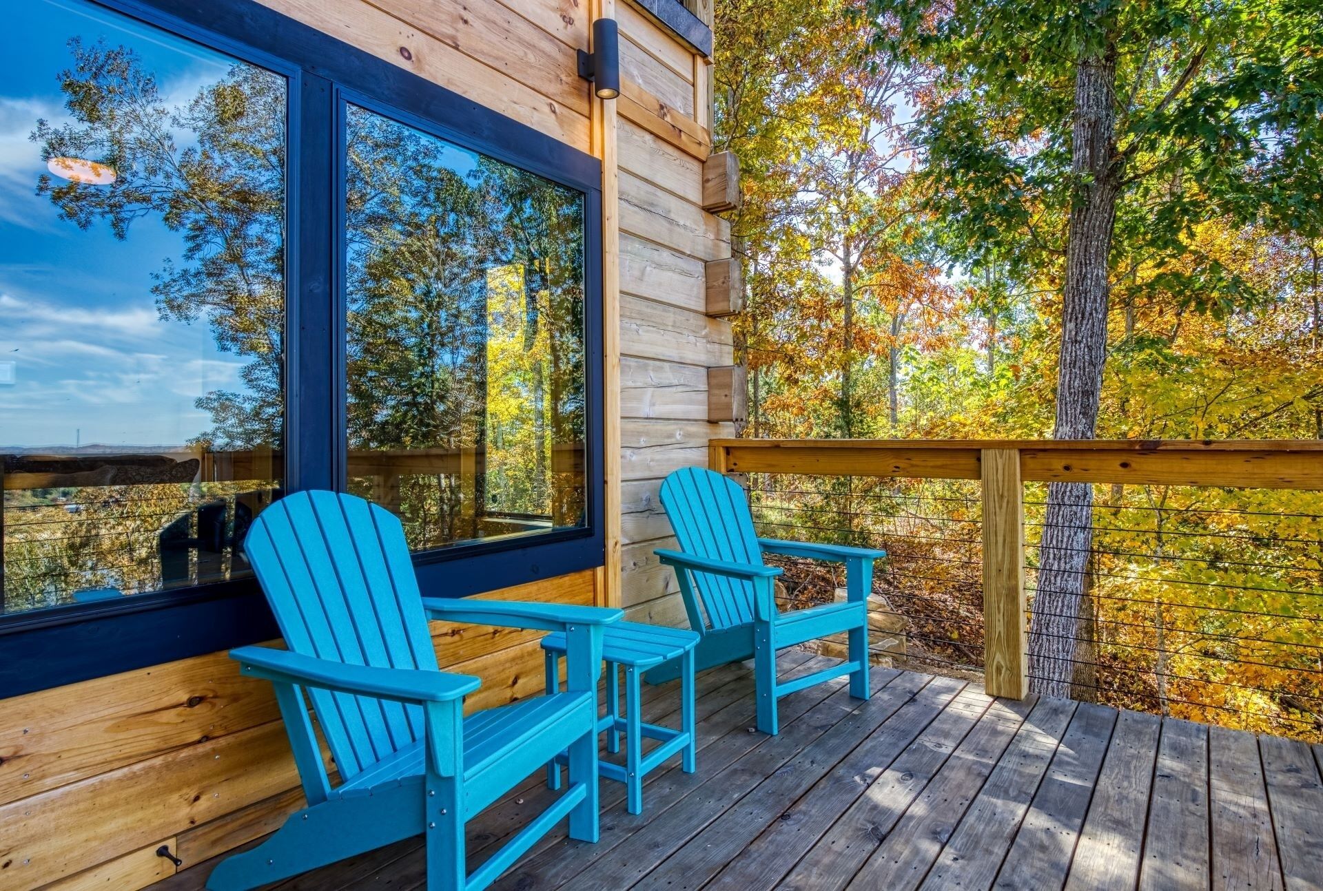 A wooden deck with two Adirondack chairs, a large window, and a view of autumn trees.