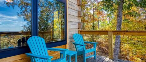 A wooden deck with two Adirondack chairs, a large window, and a view of autumn trees.