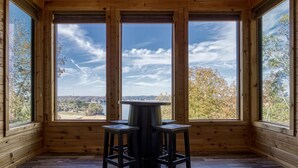 A room with wooden walls and floors, a table with two stools, and large windows offering a view of trees and a distant landscape. - Smoky Lake Retreat 3 Bedroom Cabin by RedAwning (Dandridge)
