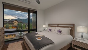 Une chambre d’hôtel dotée d’une grande fenêtre donnant sur la ville et les montagnes. La chambre comprend un lit avec du linge de lit blanc, une table de chevet avec une lampe, et une petite table sur laquelle se trouvent un croissant et un livre.