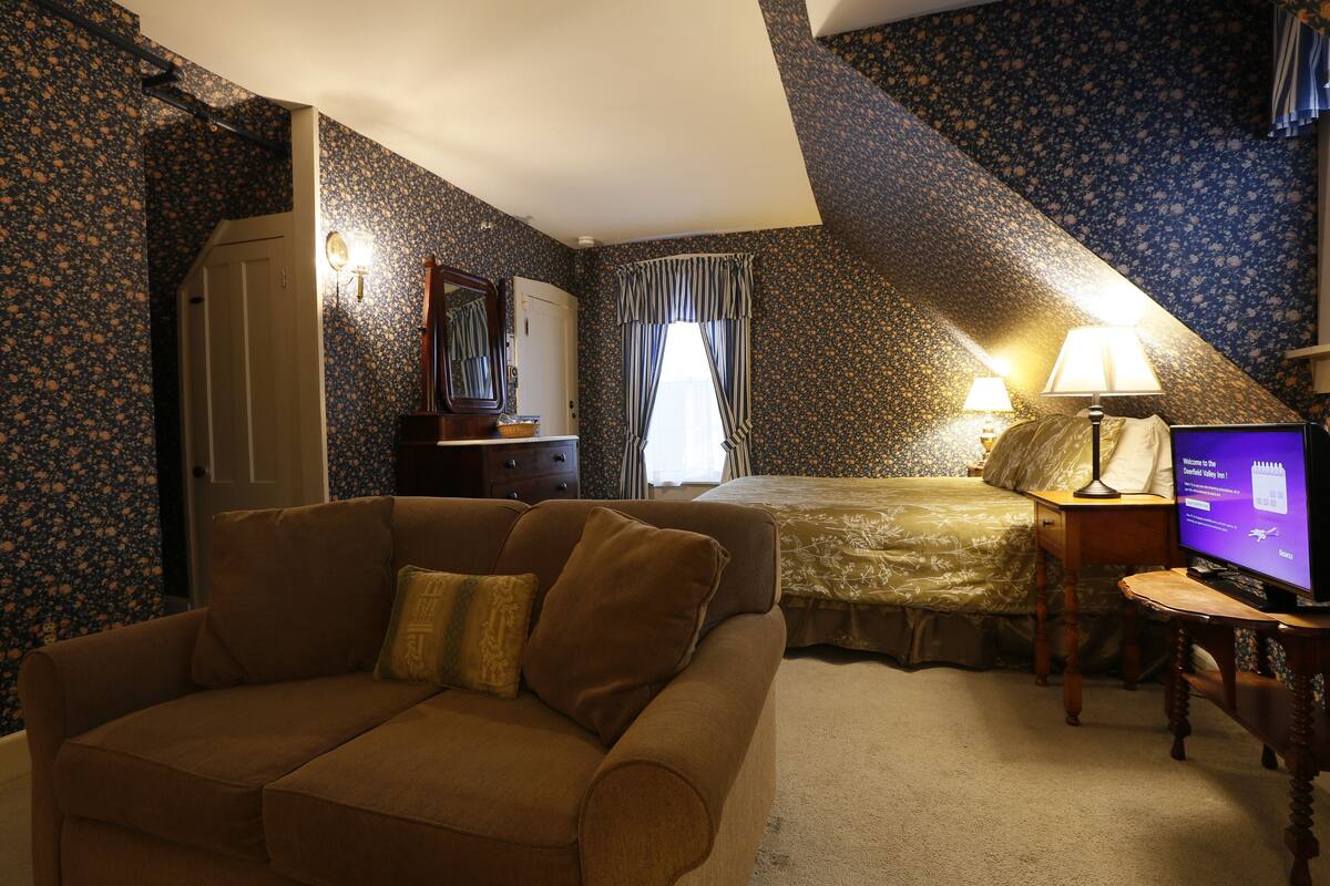 A bedroom with a sloped ceiling, a sofa, a television, a bedside table, and a bed with a floral bedspread.