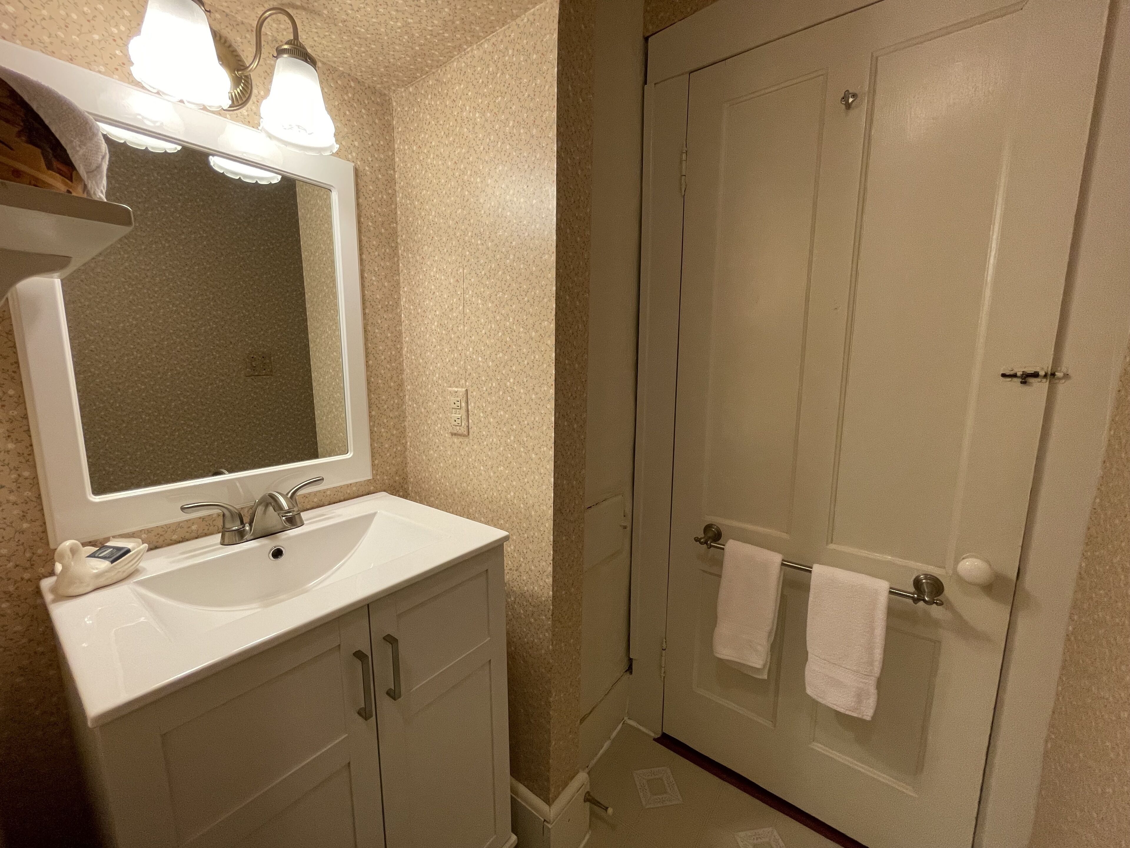 Un baño con lavamanos blanco, espejo y dos luces en el techo. Se observa una puerta con un perchero y una toalla colgando.