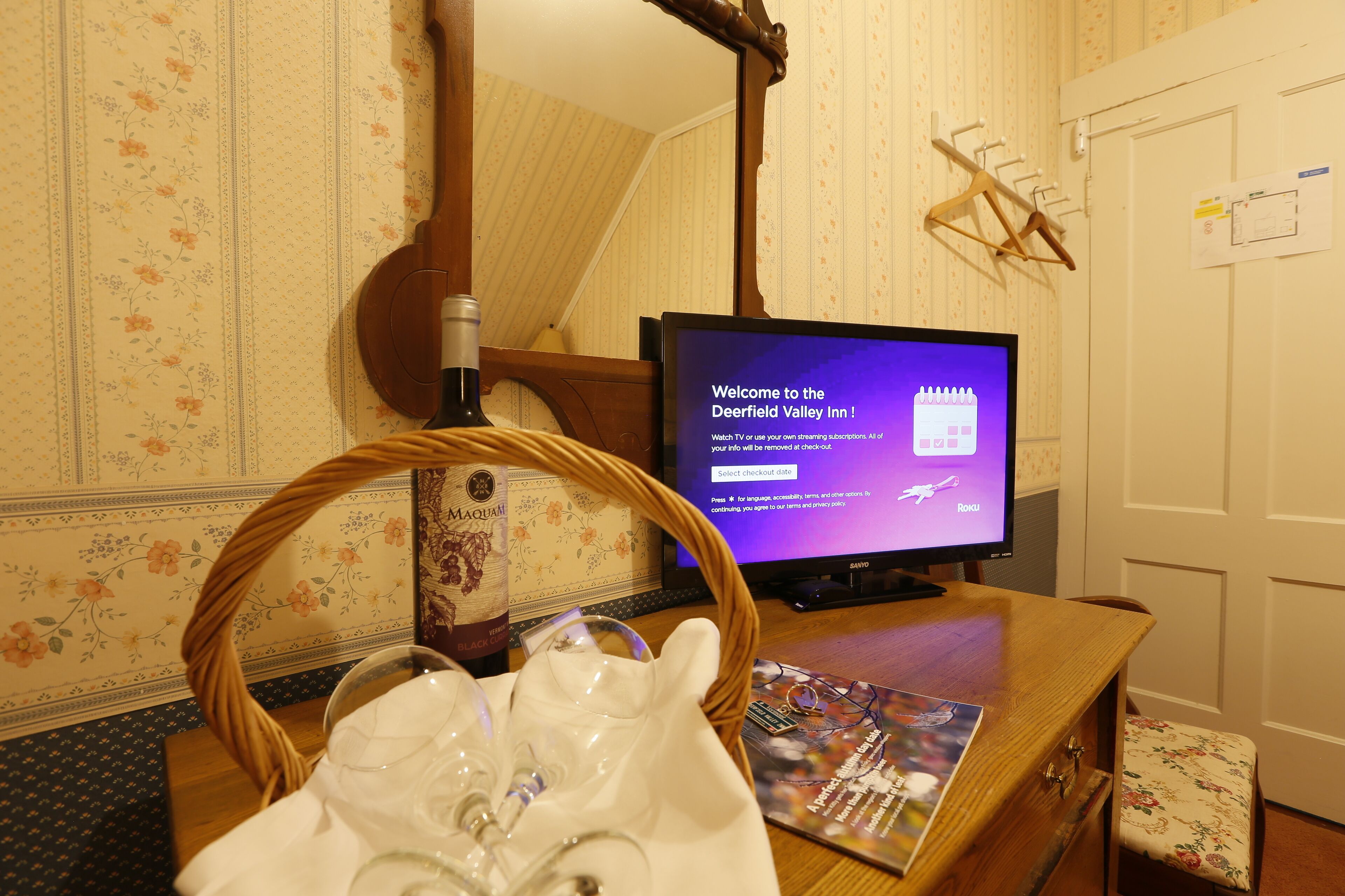 Una stanza con una televisione che mostra un messaggio di benvenuto, un cesto di vimini con una bottiglia e una rivista su un tavolo di legno.