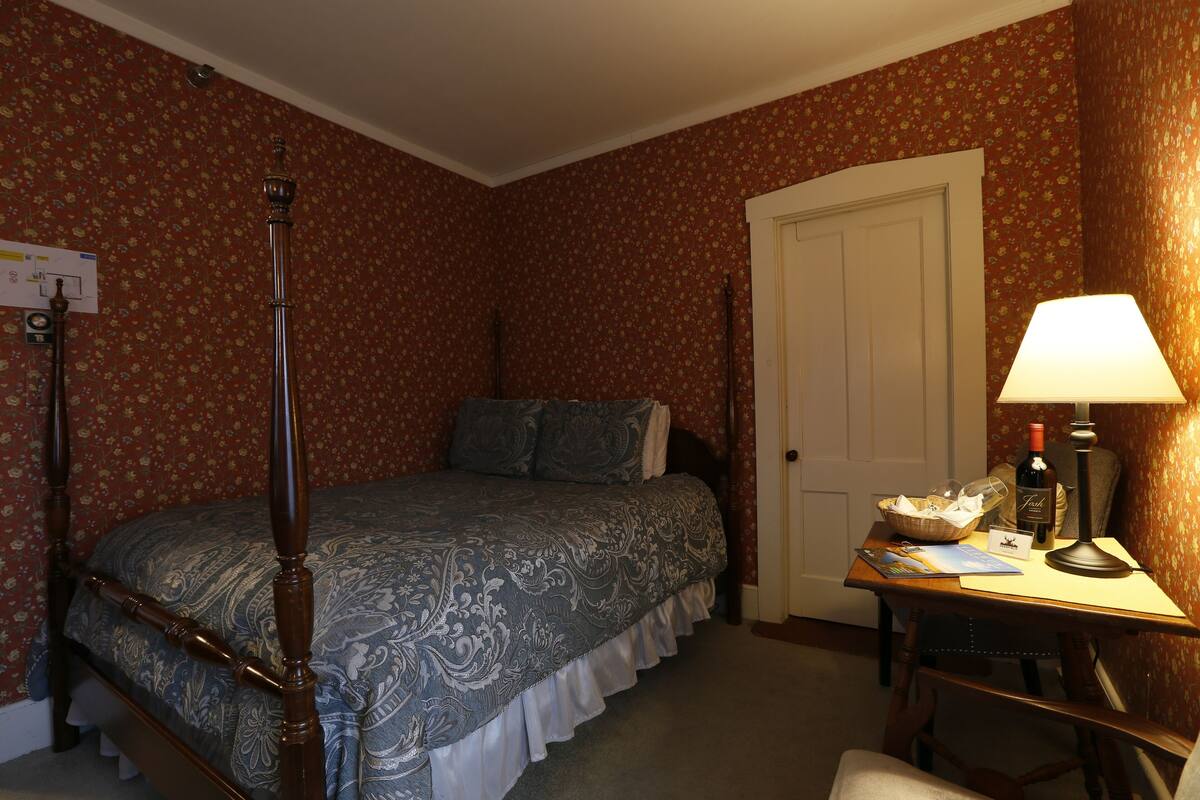 A bedroom with a four-poster bed, a desk with a lamp, and a door.