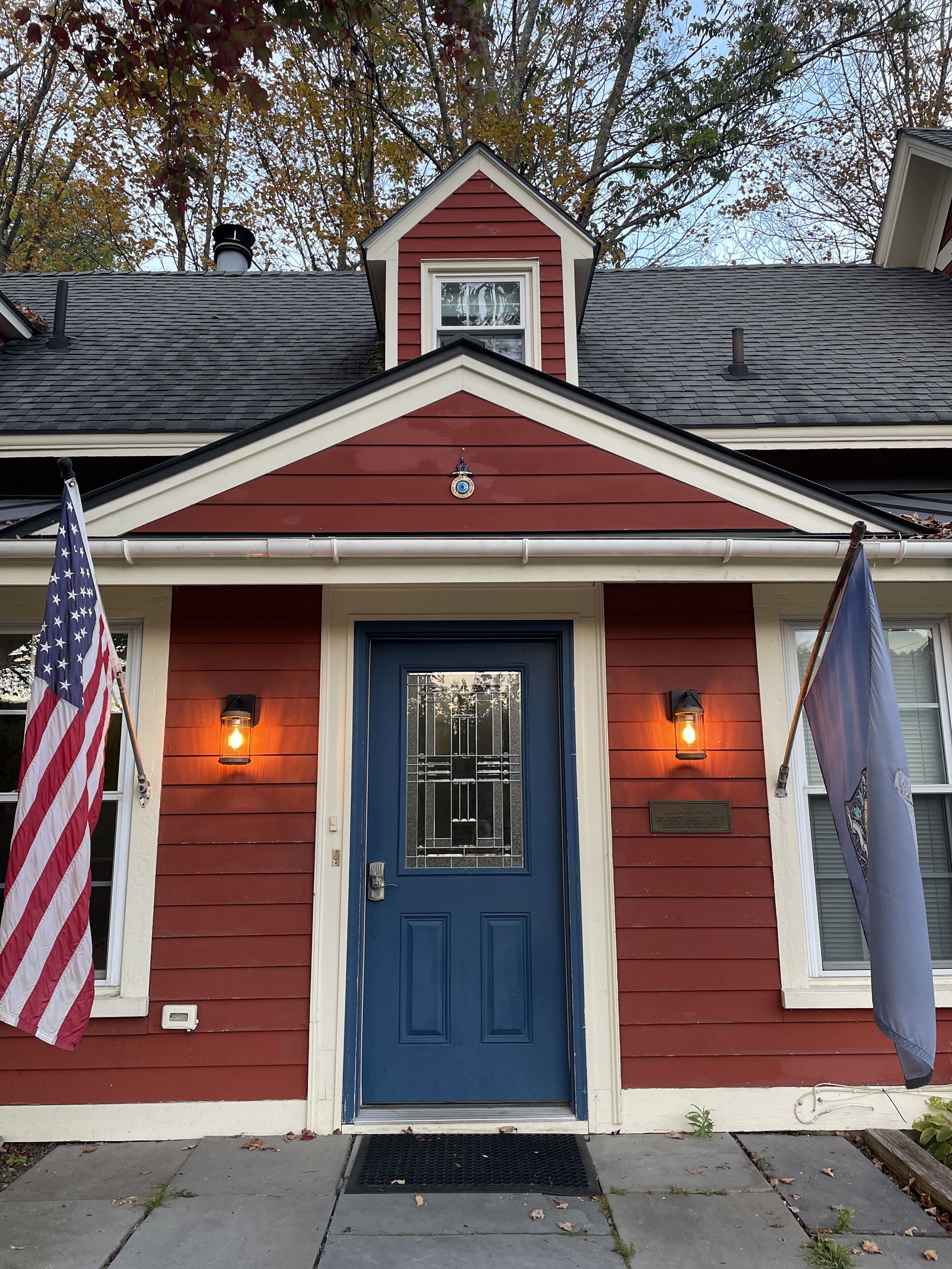 Una casa roja con una puerta azul, dos luces exteriores encendidas y una bandera estadounidense.